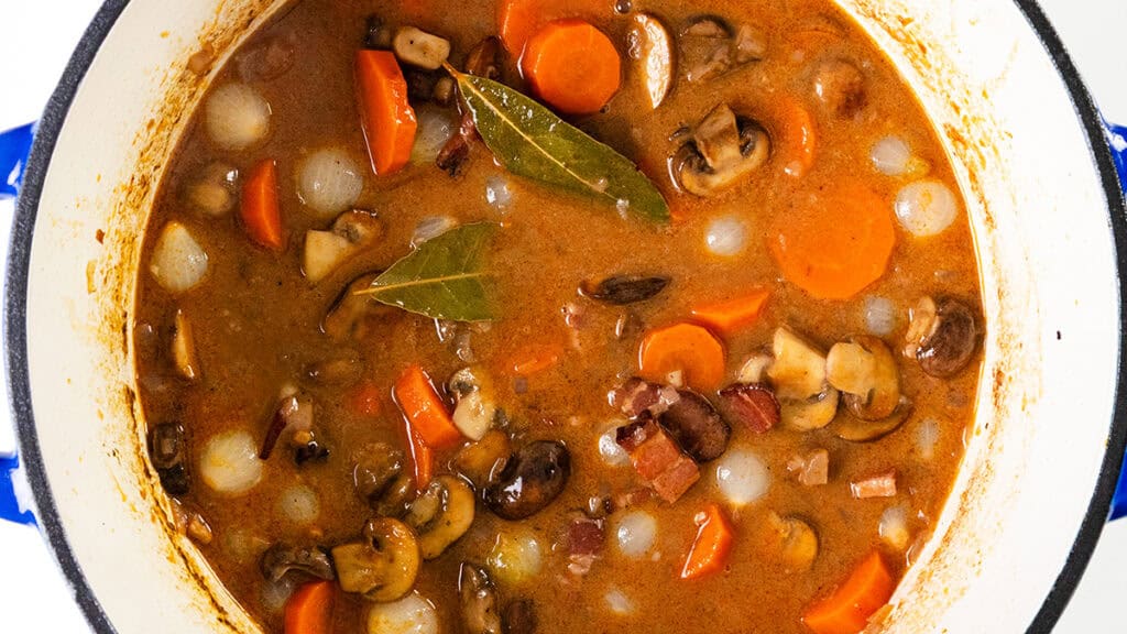 A pot of rich Coq Au Vin stew containing carrots, mushrooms, pearl onions, and bacon pieces in a savory brown broth, garnished with bay leaves. The dish appears hearty and flavorful, ideal for a comforting meal.