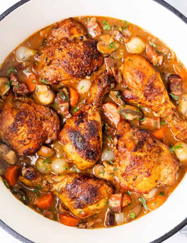 A close-up of a pot containing Coq Au Vin, showcasing succulent chicken pieces, carrots, onions, and mushrooms in a rich brown sauce. The dish is garnished with fresh herbs, creating an appetizing view of this classic hearty meal.