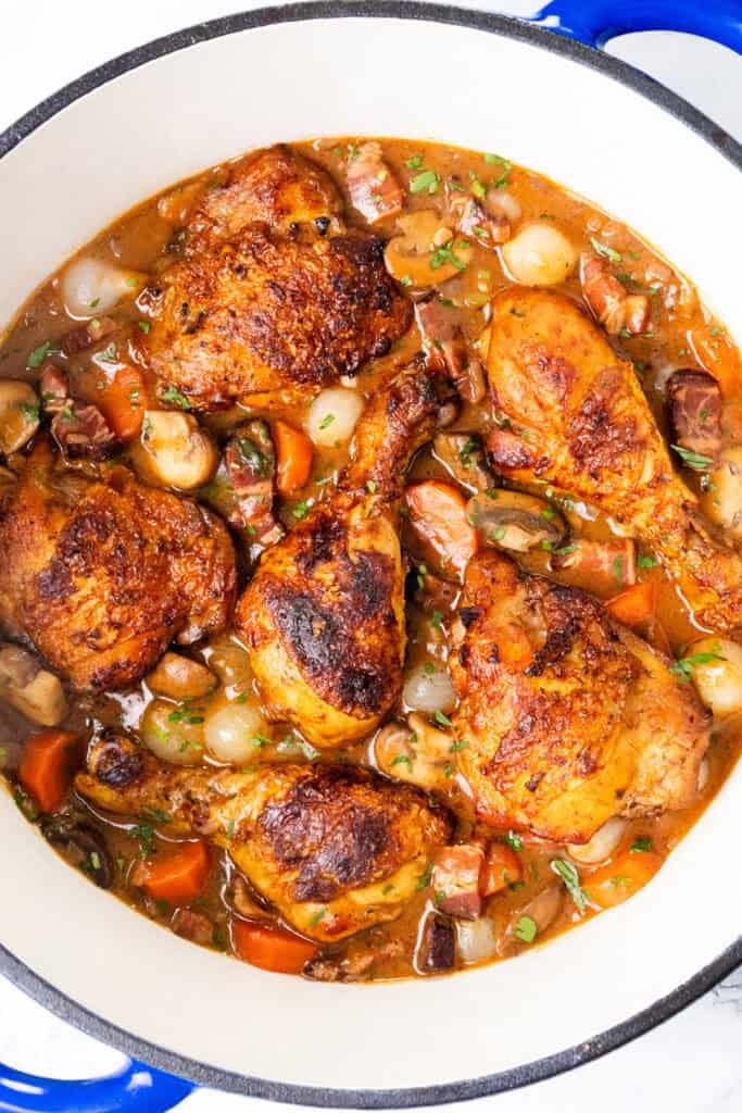 A close-up of a pot containing Coq Au Vin, showcasing succulent chicken pieces, carrots, onions, and mushrooms in a rich brown sauce. The dish is garnished with fresh herbs, creating an appetizing view of this classic hearty meal.