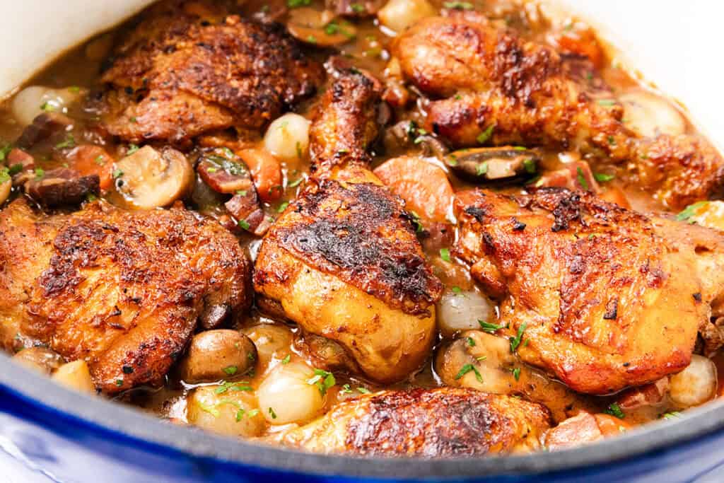 A close-up of a Coq Au Vin-inspired dish featuring browned chicken thighs with mushrooms, carrots, and pearl onions in a savory sauce, garnished with chopped herbs, all simmering deliciously in a pot.