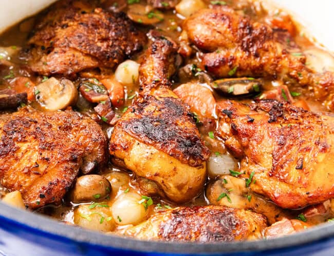 A close-up of a Coq Au Vin-inspired dish featuring browned chicken thighs with mushrooms, carrots, and pearl onions in a savory sauce, garnished with chopped herbs, all simmering deliciously in a pot.