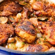 A close-up of a pot filled with golden-brown roasted chicken pieces, mushrooms, carrots, onions, and herbs in a savory Coq Au Vin sauce.