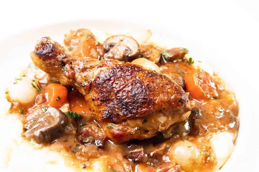 A savory chicken drumstick reminiscent of Coq Au Vin sits atop a bed of creamy mashed potatoes, surrounded by a rich stew of mushrooms, carrots, onions, and herbs on a white plate.