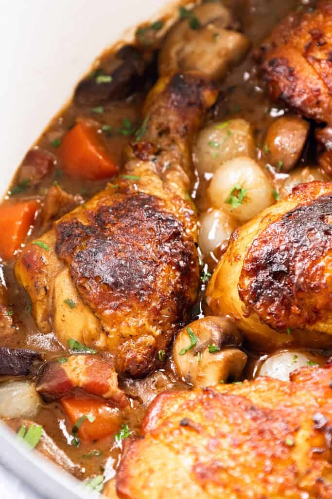 Close-up of a Coq Au Vin-inspired dish featuring roasted chicken legs with golden-brown skin, surrounded by carrots, pearl onions, and mushrooms in a rich, savory sauce. The dish is garnished with chopped herbs.