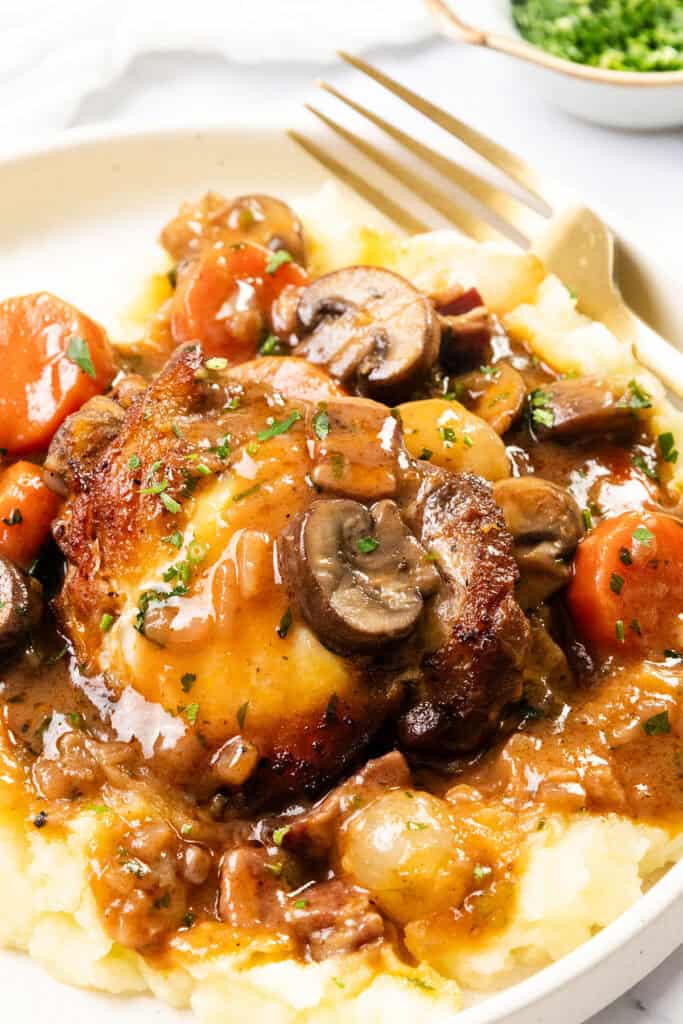 A plate of mashed potatoes topped with a richly sauced chicken thigh, mushrooms, and carrots reminiscent of Coq Au Vin. Garnished with chopped herbs, the dish is surrounded by a fork and a blurred bowl of green garnish in the background.