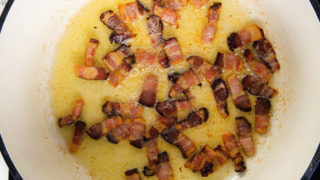 Crispy diced bacon frying in a pan, surrounded by melted fat, reminiscent of the rich flavors found in a classic Coq Au Vin. The golden-brown pieces with darker edges rest against the light-colored backdrop of the pan.