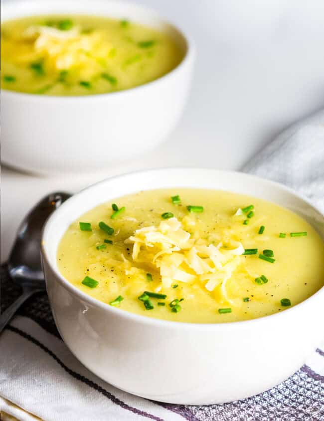 A white bowl filled with creamy potato leek soup topped with shredded cheese and chopped green onions, sitting on a folded striped cloth with a spoon nearby. Another bowl of soup is blurred in the background.