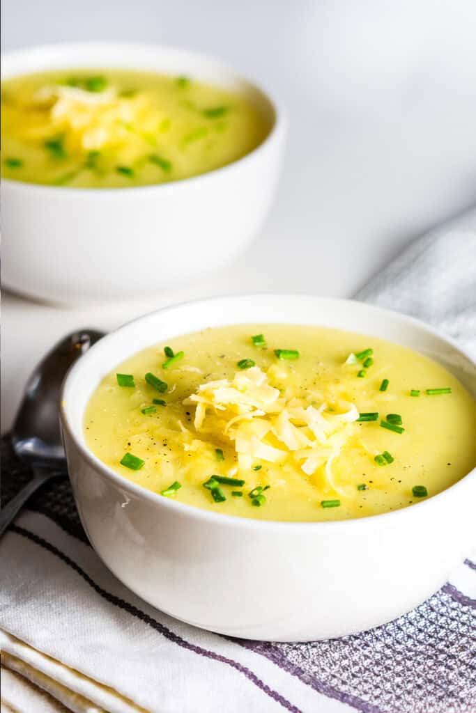 A white bowl filled with creamy potato leek soup topped with shredded cheese and chopped green onions, sitting on a folded striped cloth with a spoon nearby. Another bowl of soup is blurred in the background.