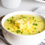 A white bowl filled with creamy potato leek soup, topped with shredded cheese and chopped chives. A spoon lies next to the bowl on a striped cloth napkin. In the background, there's a white dish and another bowl, partially visible.