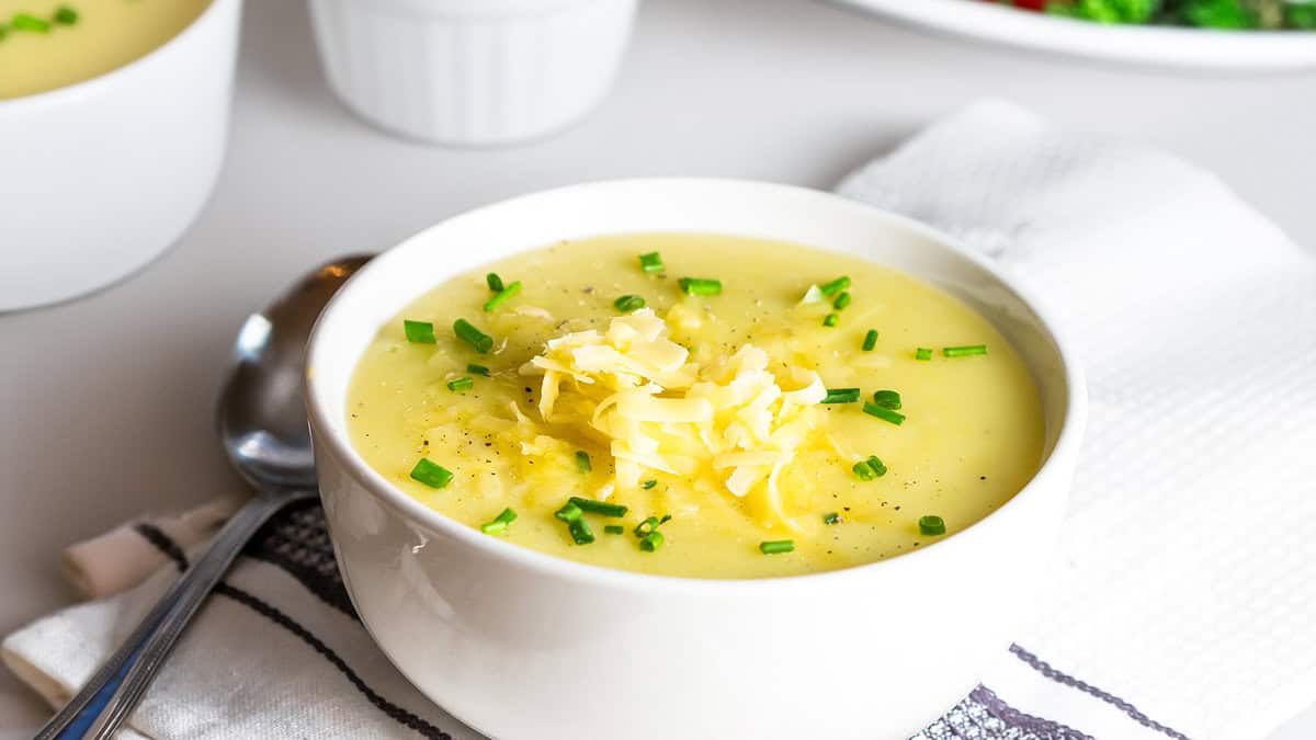 A white bowl filled with creamy potato leek soup, topped with shredded cheese and chopped chives. A spoon lies next to the bowl on a striped cloth napkin. In the background, there's a white dish and another bowl, partially visible.