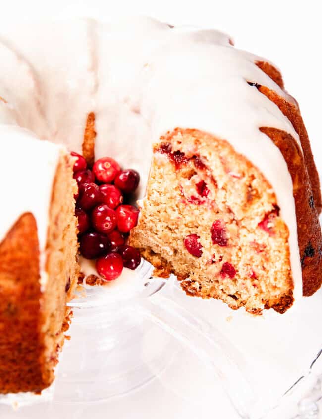 A Cranberry Christmas Cake with sliced portions, topped with white glaze and garnished with red cranberries, displayed on a clear glass stand. A section of this festive cake is cut to reveal a moist interior dotted with juicy cranberries.