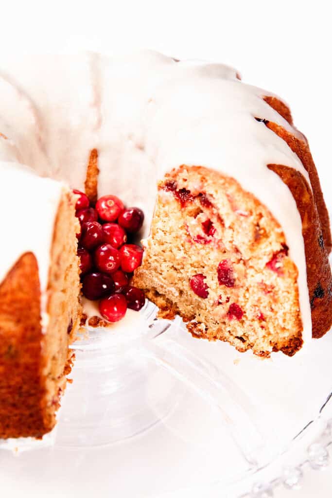 A Cranberry Christmas Cake with sliced portions, topped with white glaze and garnished with red cranberries, displayed on a clear glass stand. A section of this festive cake is cut to reveal a moist interior dotted with juicy cranberries.