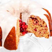 A Cranberry Christmas Bundt cake with a slice removed is displayed on a glass stand. The cake, topped with white icing, reveals a texture speckled with red berries inside. A cluster of fresh red berries elegantly decorates the center.