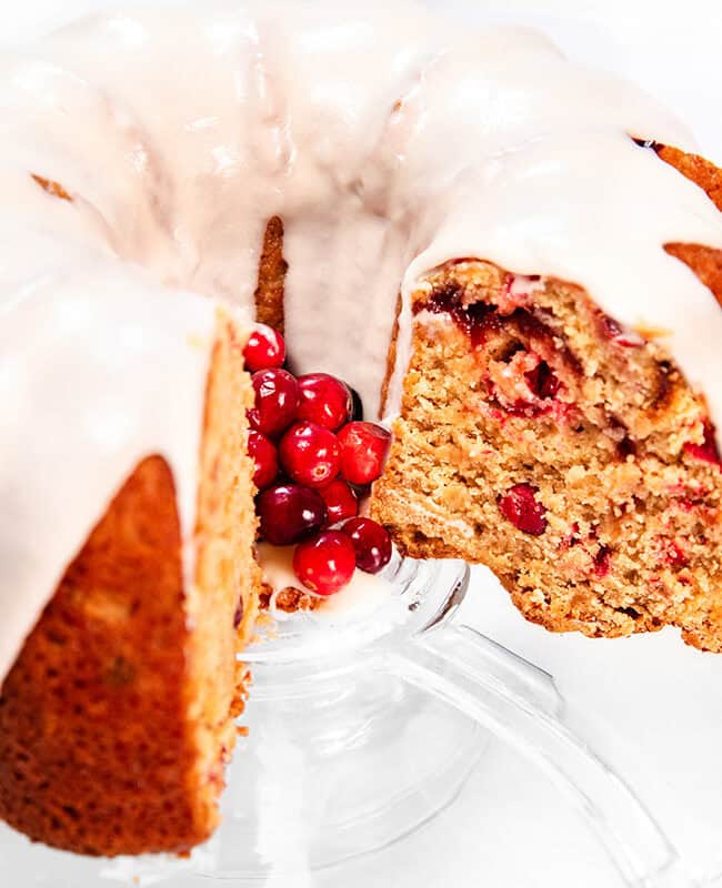 A Cranberry Christmas Bundt cake with a slice removed is displayed on a glass stand. The cake, topped with white icing, reveals a texture speckled with red berries inside. A cluster of fresh red berries elegantly decorates the center.