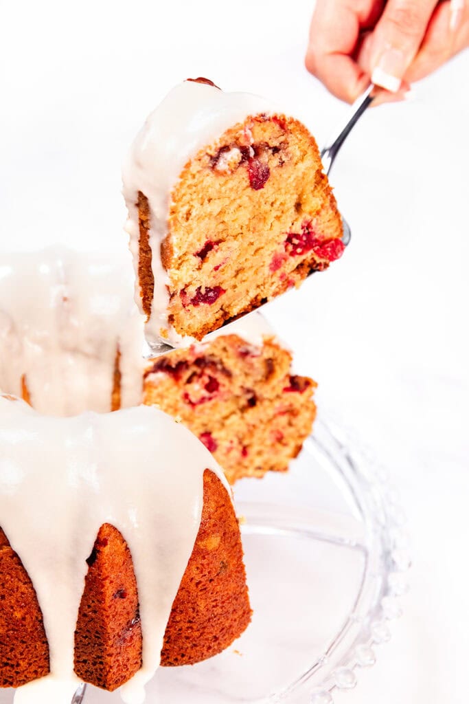 A hand lifts a slice of Cranberry Christmas Cake with white icing from a glass cake stand. The festive bundt cake is filled with pieces of red fruit, possibly cranberries, making it perfect for holiday celebrations.