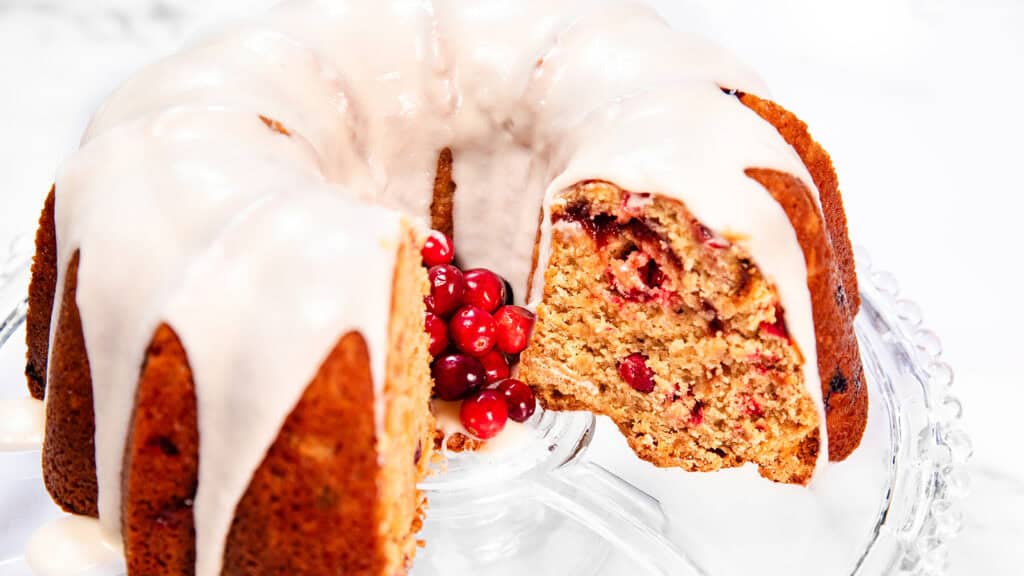 A Cranberry Christmas Cake with white icing sits elegantly on a glass cake stand. A slice is cut out, revealing its moist interior speckled with red berries. A festive cluster of cranberries adorns the center, completing this holiday delight.