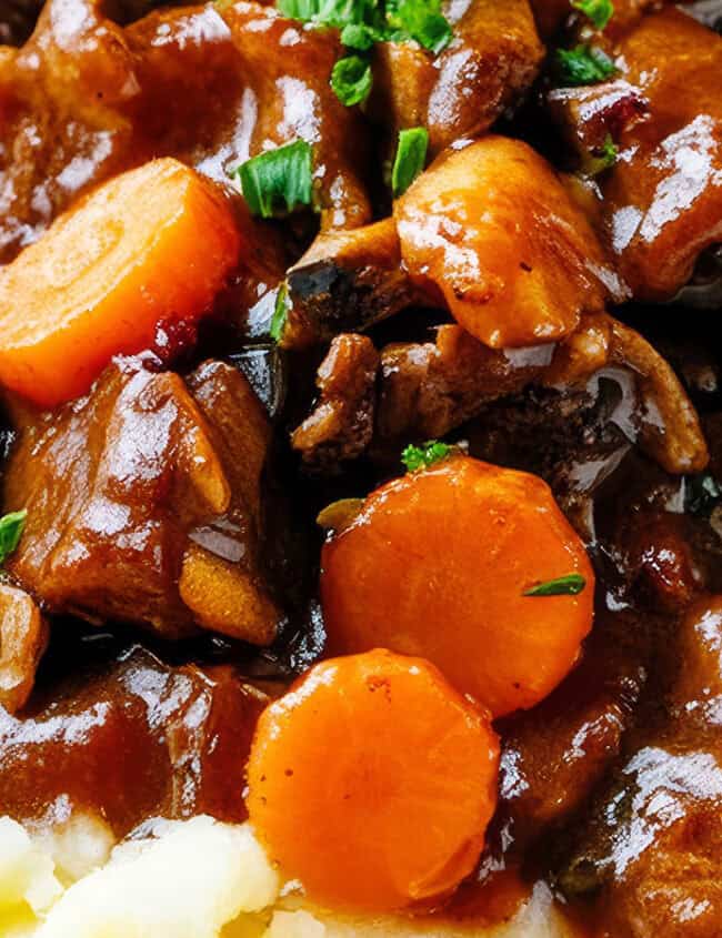 Close-up of a hearty beef stew with chunks of tender beef, carrots, and mushrooms covered in rich gravy, served on a bed of creamy mashed potatoes. The dish features braised boneless short ribs and is garnished with finely chopped fresh herbs.