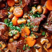 A hearty beef stew in a pot, featuring chunks of tender beef and braised short ribs, sliced carrots, whole mushrooms, and garnished with chopped fresh parsley. The rich, dark broth adds a comforting warmth to the beautifully presented dish.