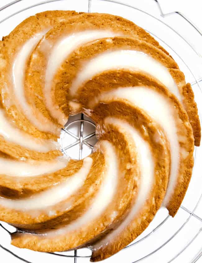 A glazed bundt cake with spiral ridges sits on a round cooling rack. This easy banana cake flaunts a golden brown hue with a shiny white glaze that fills the grooves, creating a striped pattern. The background is white, emphasizing the cake's texture and design.