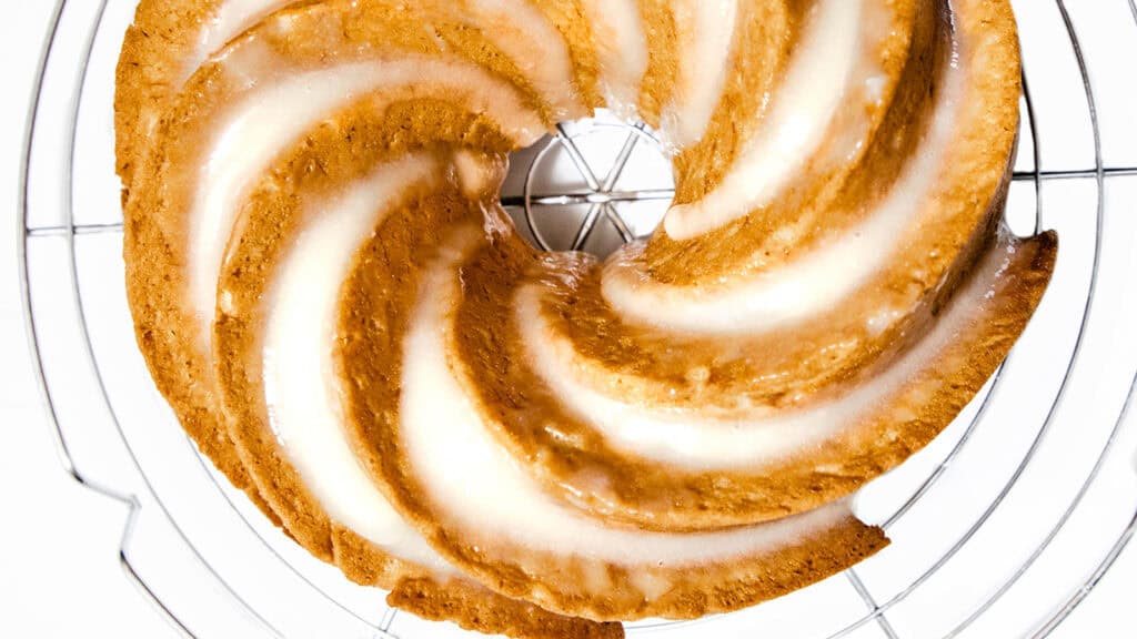 A beautifully baked bundt cake with a white glaze sits on a wire cooling rack. This easy banana cake has a spiral pattern and a golden brown hue, with the glossy glaze accentuating its delicate texture.