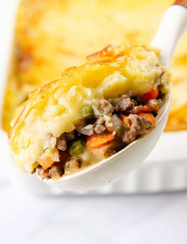 A spoonful of cottage pie is being lifted above a baking dish. The pie boasts a golden-brown mashed potato topping, with a savory filling of ground meat, peas, carrots, and onions. In the background, the rest of the deliciously baked dish awaits.