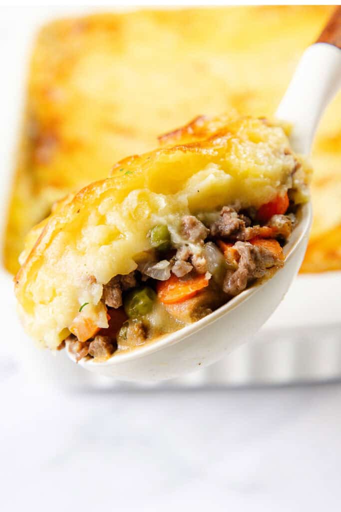 A spoonful of cottage pie is being lifted above a baking dish. The pie boasts a golden-brown mashed potato topping, with a savory filling of ground meat, peas, carrots, and onions. In the background, the rest of the deliciously baked dish awaits.