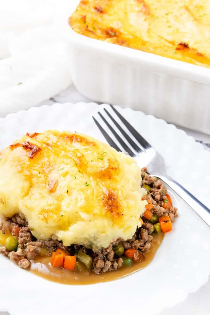 A serving of cottage pie on a white plate, showcasing perfectly browned mashed potatoes atop a savory filling of minced meat and mixed vegetables like peas and carrots. A fork rests beside the dish, with a casserole dish in the background.