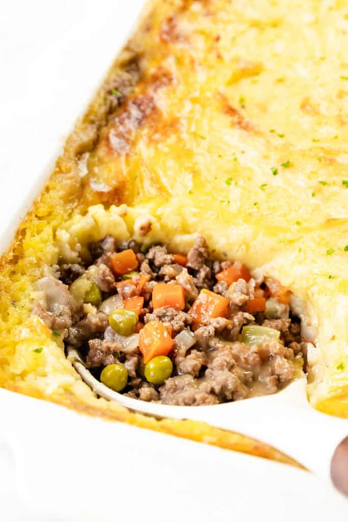 A close-up of a dish of cottage pie, showcasing the savory minced meat filling with peas, carrots, and onions beneath a layer of creamy mashed potatoes. A portion is scooped out, revealing the vibrant layers in this comforting classic.