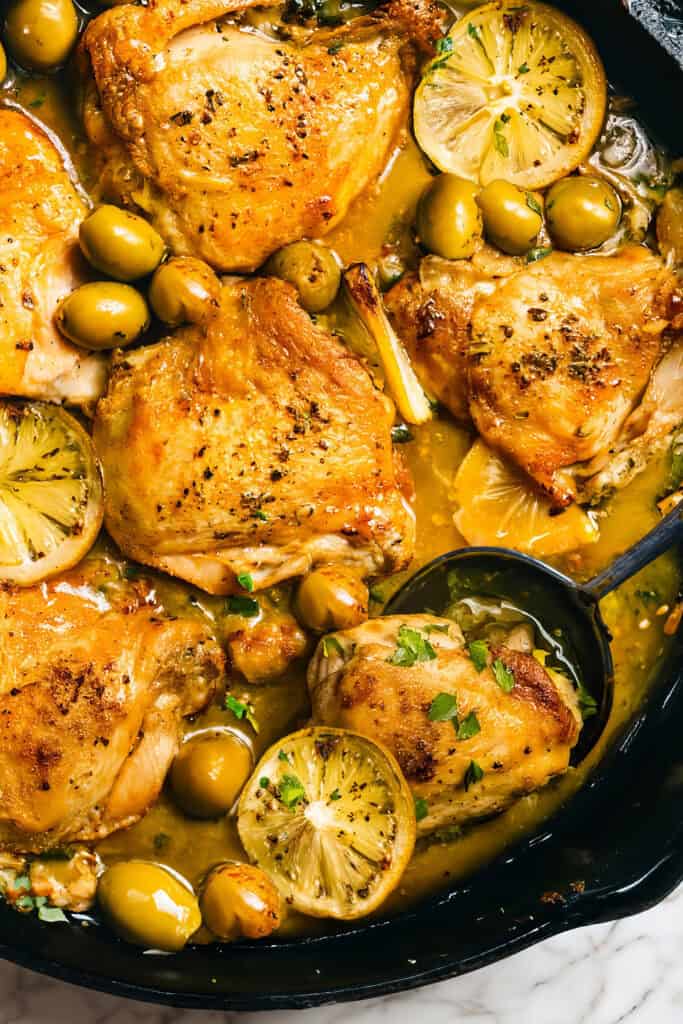 A skillet brimming with Moroccan chicken thighs, golden-brown and garnished with lemon slices, green olives, and fresh herbs. The dish is enveloped in a savory sauce, with a spoon dipping into the aromatic blend.