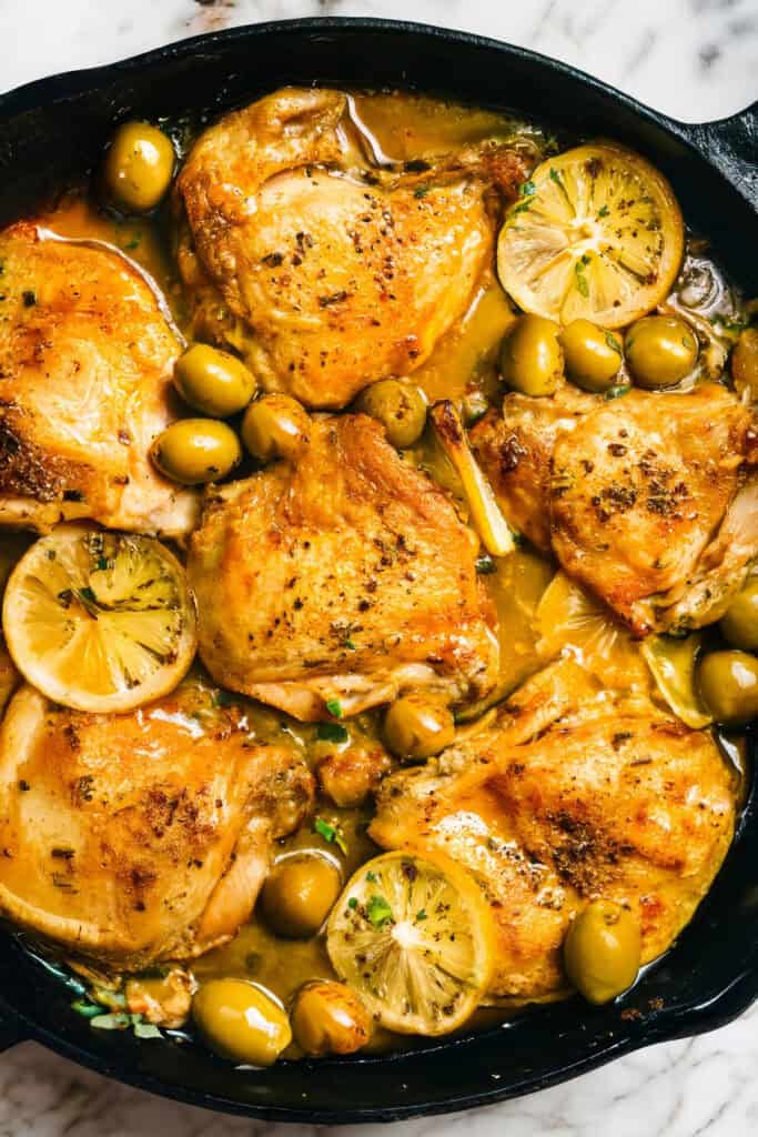 A skillet filled with golden-brown Moroccan chicken thighs, sliced lemons, and green olives, garnished with fresh thyme.