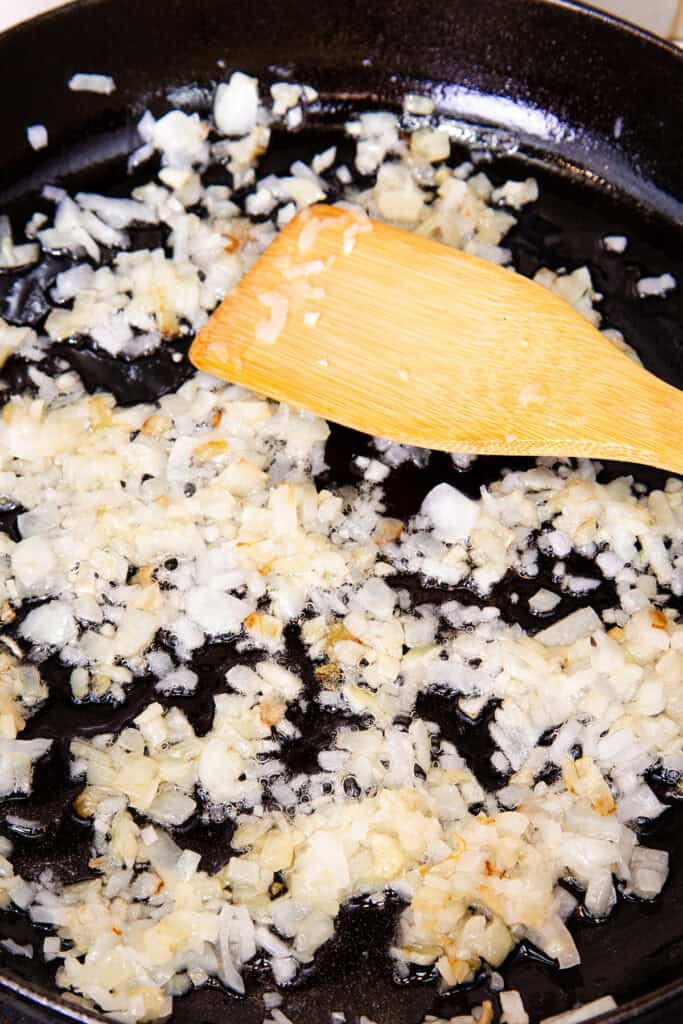 Chopped onions being sautéed in a black skillet with a wooden spatula, the fragrant base for Moroccan chicken.