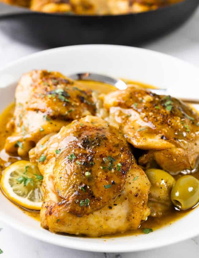 A plate of Moroccan chicken thighs garnished with chopped herbs awaits. The dish features lemon slices and olives, resting on a white plate. In the background, a cast-iron skillet holds more flavorful chicken, promising a delectable feast.