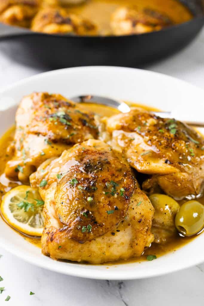 A plate of Moroccan chicken thighs garnished with chopped herbs awaits. The dish features lemon slices and olives, resting on a white plate. In the background, a cast-iron skillet holds more flavorful chicken, promising a delectable feast.