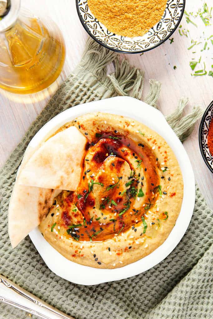 A bowl of Baba Ghanouj garnished with fresh herbs, served with pita bread, olive oil, and spices on a textured green cloth.