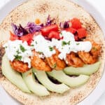 A shrimp taco on a toasted tortilla with avocado slices, red cabbage, diced tomato, and a creamy white sauce, garnished with chopped herbs on a white plate.