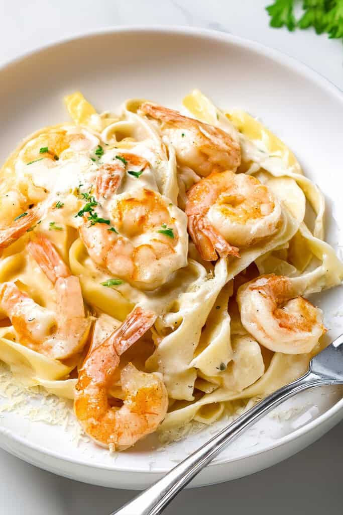 A white bowl filled with succulent Shrimp Alfredo Pasta, featuring fettuccine coated in creamy Alfredo sauce, topped with perfectly grilled shrimp and garnished with chopped parsley. A silver fork rests on the side of the bowl.