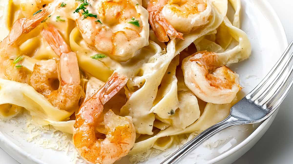 A close-up of a plate of Shrimp Alfredo Pasta. The pasta appears to be coated in a creamy white sauce, and the shrimp are large and pink, garnished with a sprinkle of herbs. A fork rests on the plate's rim.