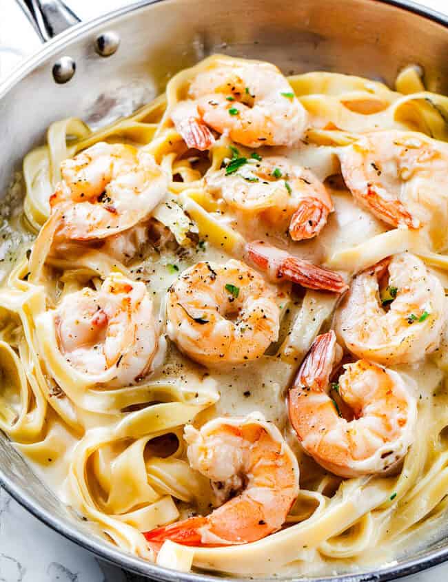 A close-up of a skillet containing creamy Shrimp Alfredo Pasta. The dish features fettuccine noodles in a rich, creamy sauce, topped with plump, cooked shrimp. The shrimp are garnished with herbs and black pepper, and the pasta is well-coated with the creamy sauce.