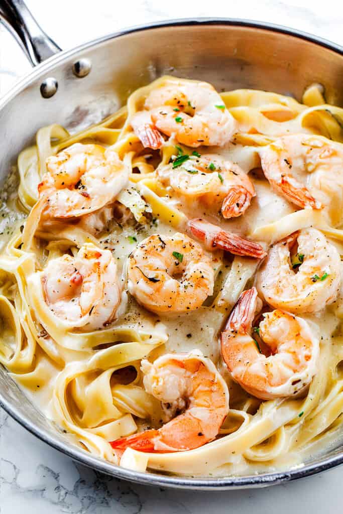 A close-up of a skillet containing creamy Shrimp Alfredo Pasta. The dish features fettuccine noodles in a rich, creamy sauce, topped with plump, cooked shrimp. The shrimp are garnished with herbs and black pepper, and the pasta is well-coated with the creamy sauce.