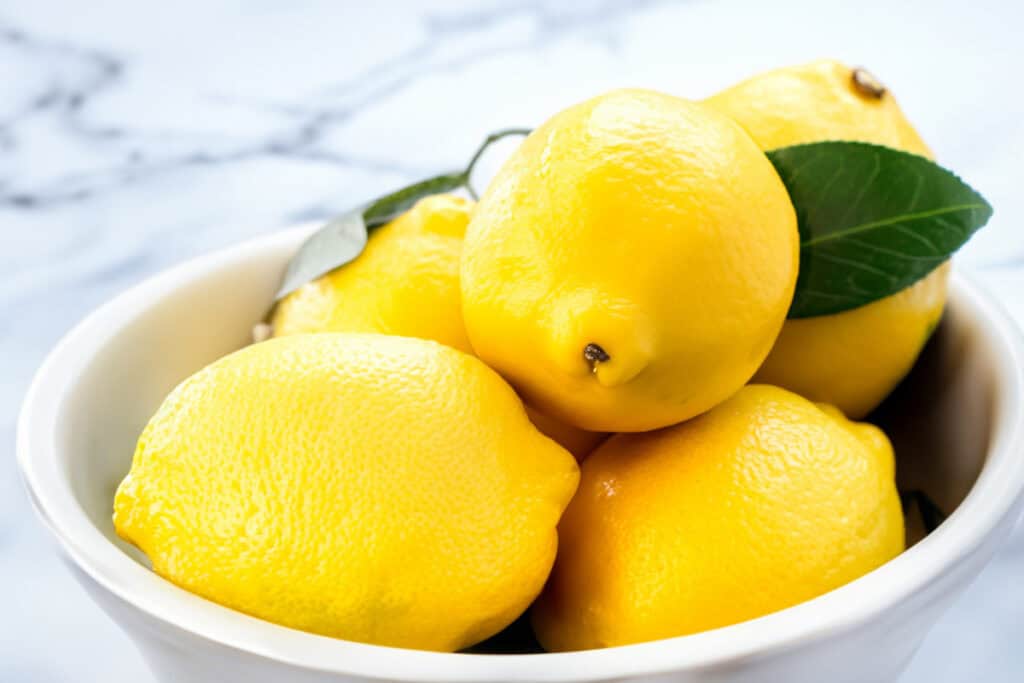 A close up image of a bowl of lemons on a white marble surface.