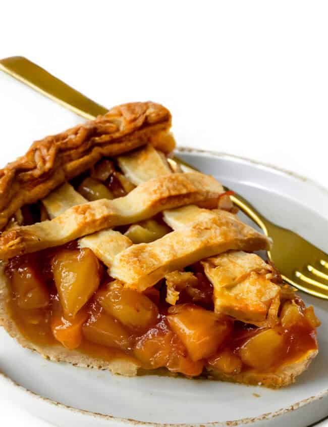 A slice of peach pie with a golden-brown lattice crust is placed on a white plate. The vibrant peach chunks in the filling evoke the charm of an Old Fashioned Apple Spice Pie. A gold-colored fork is partially visible beneath the pie on the plate.