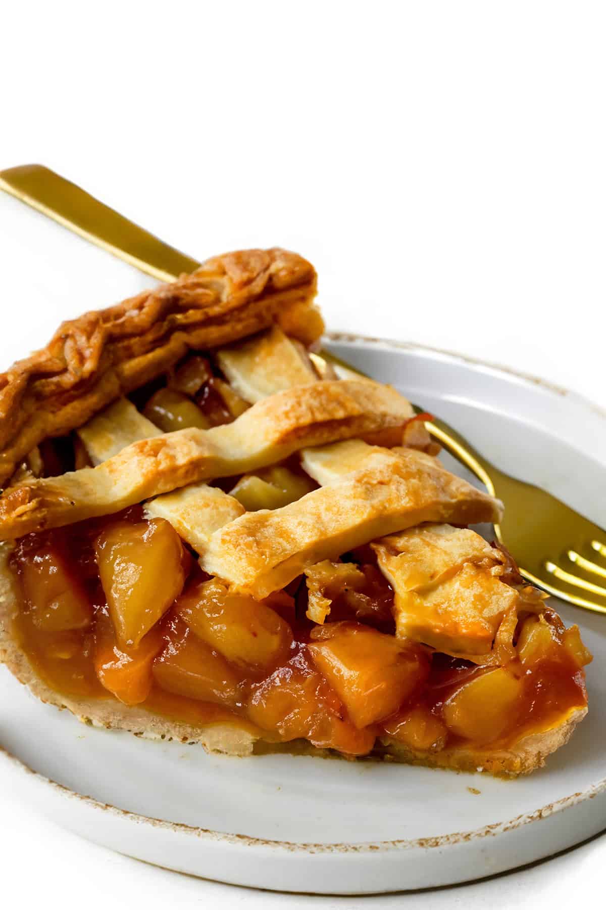 A slice of peach pie with a golden-brown lattice crust is placed on a white plate. The vibrant peach chunks in the filling evoke the charm of an Old Fashioned Apple Spice Pie. A gold-colored fork is partially visible beneath the pie on the plate.