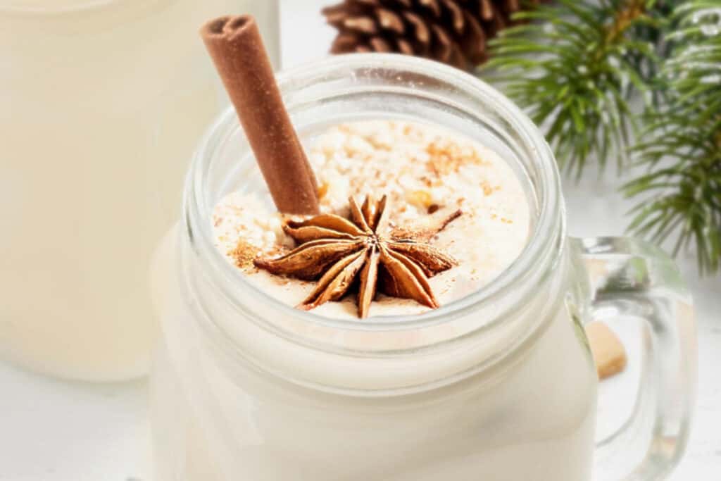 a mug of fresh eggnog garnished with star anise and a cinnamon stick with Christmas garland in the background