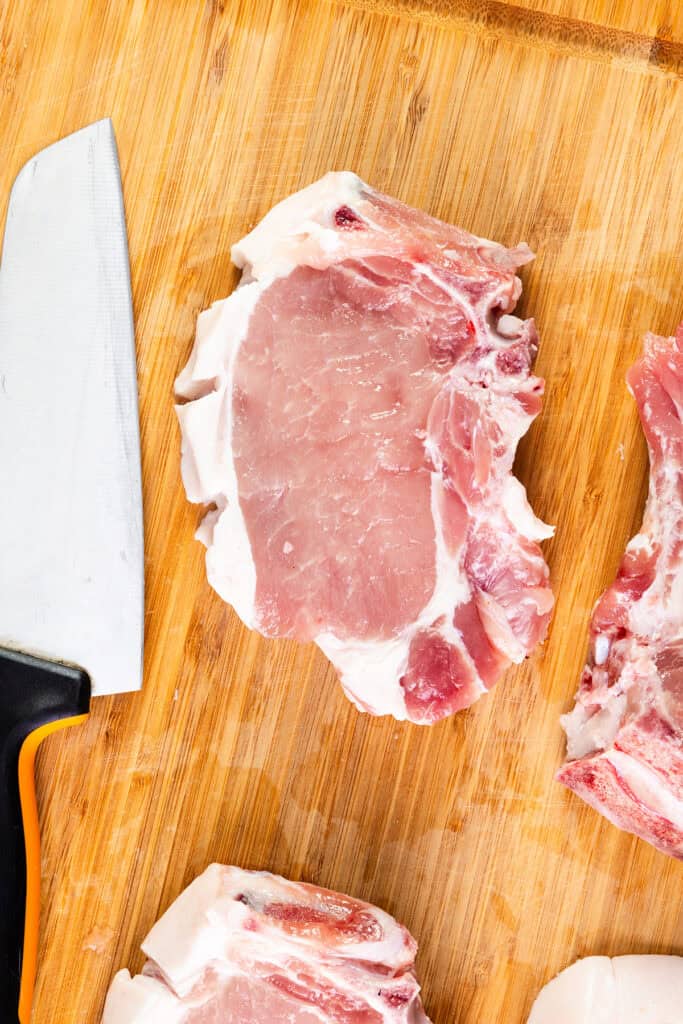 A raw brined pork chop, partially cut, sits on a wooden cutting board with a kitchen knife visible on the left side. Additional pieces of meat are seen in the lower part of the image, ready to be pan seared for a delicious meal.