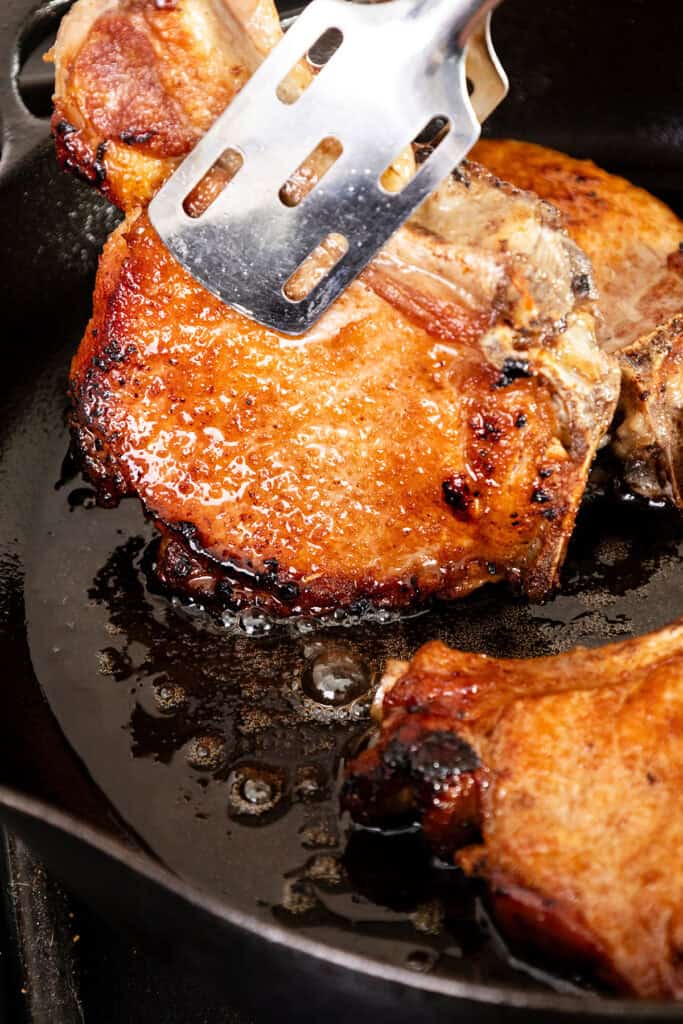 A metal spatula presses down on a juicy, golden-brown brined pork chop sizzling in a cast-iron skillet. The meat appears perfectly seared, with a slight char, and the glistening surface indicates it is being cooked in its own juices and oils.