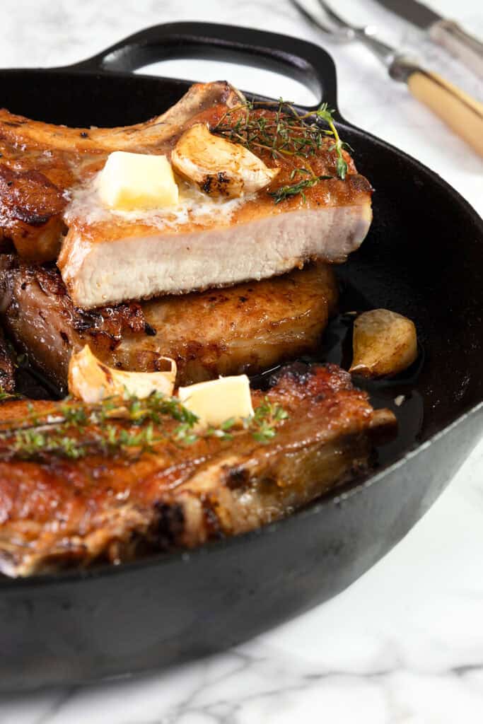 A close-up of a black cast-iron skillet containing several pan-seared brined pork chops, garnished with fresh herbs, garlic cloves, and melting pats of butter. The textured, golden-brown sear on the pork chops is visible, and cutlery rests on the marble surface beside the skillet.