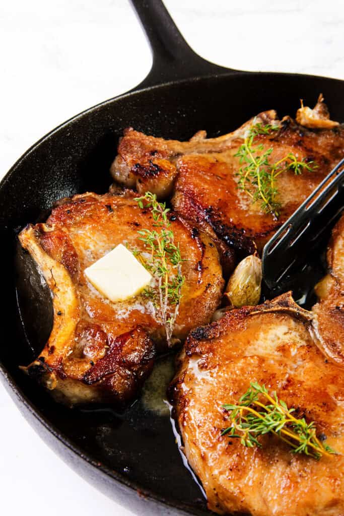 A close-up of a cast iron pan containing three pan-seared brined pork chops garnished with fresh herbs and a small pat of butter on top. The pork chops are golden brown and cooked to perfection. The pan is placed on a white marble surface.