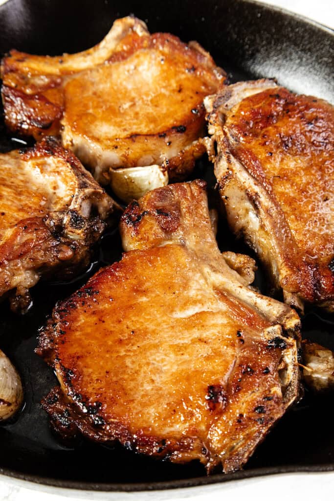 A close-up of golden brown pan seared brined pork chops frying in a black skillet. The meat is perfectly seared and appears juicy with a crispy exterior. Small bits of garlic can be seen among the pork chops, adding texture and flavor to the scene.