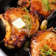 A close-up shot of a skillet with perfectly pan seared brined pork chops, garnished with sprigs of fresh thyme. A small pat of butter is melting on one of the chops. The skillet is filled with rich juices, highlighting the brined pork chops' caramelized surfaces.