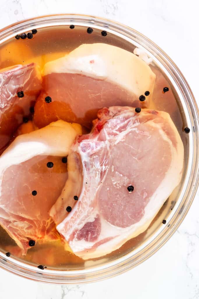 A bowl filled with pork chops soaked in brine, peppercorns scattered around. These raw pork chops are submerged in a marinating mixture, preparing them to become juicy Pan Seared Brined Pork Chops. The background features a light-colored countertop.